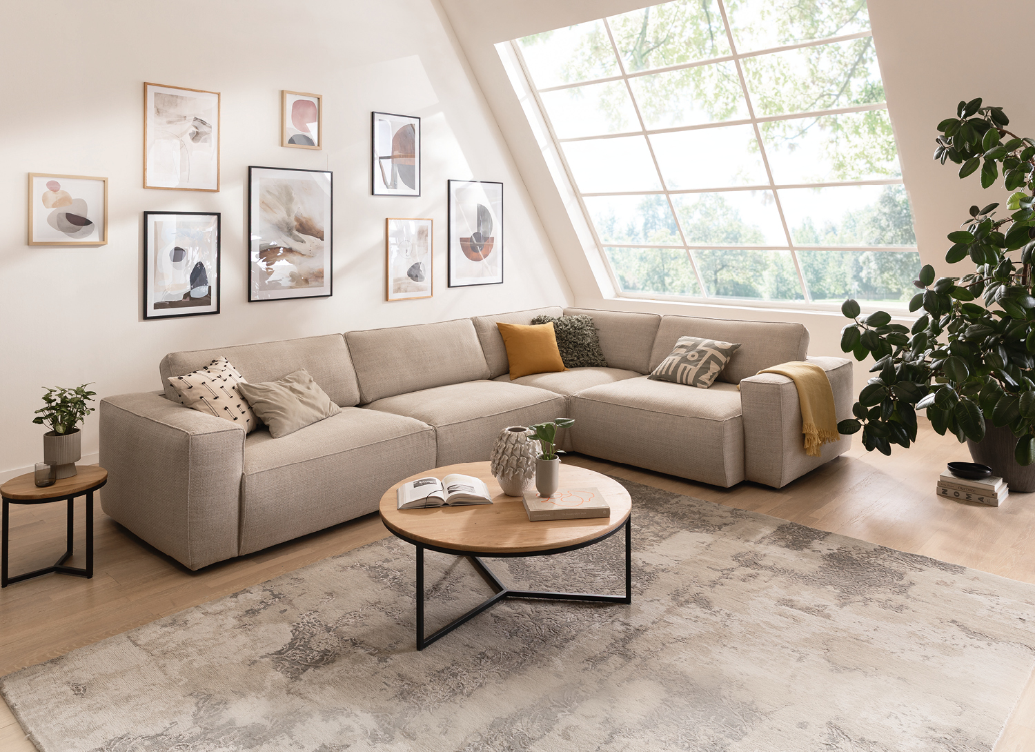 Helles Ecksofa mit rundem Couchtisch, Teppich, Bilderwand und großer Pflanze am Fenster