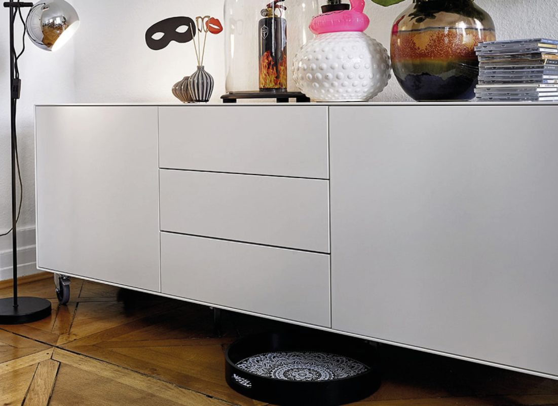 Weißes Sideboard mit Lampe, Vasen, Glasobjekten und CD Stapeln auf Holzfußboden