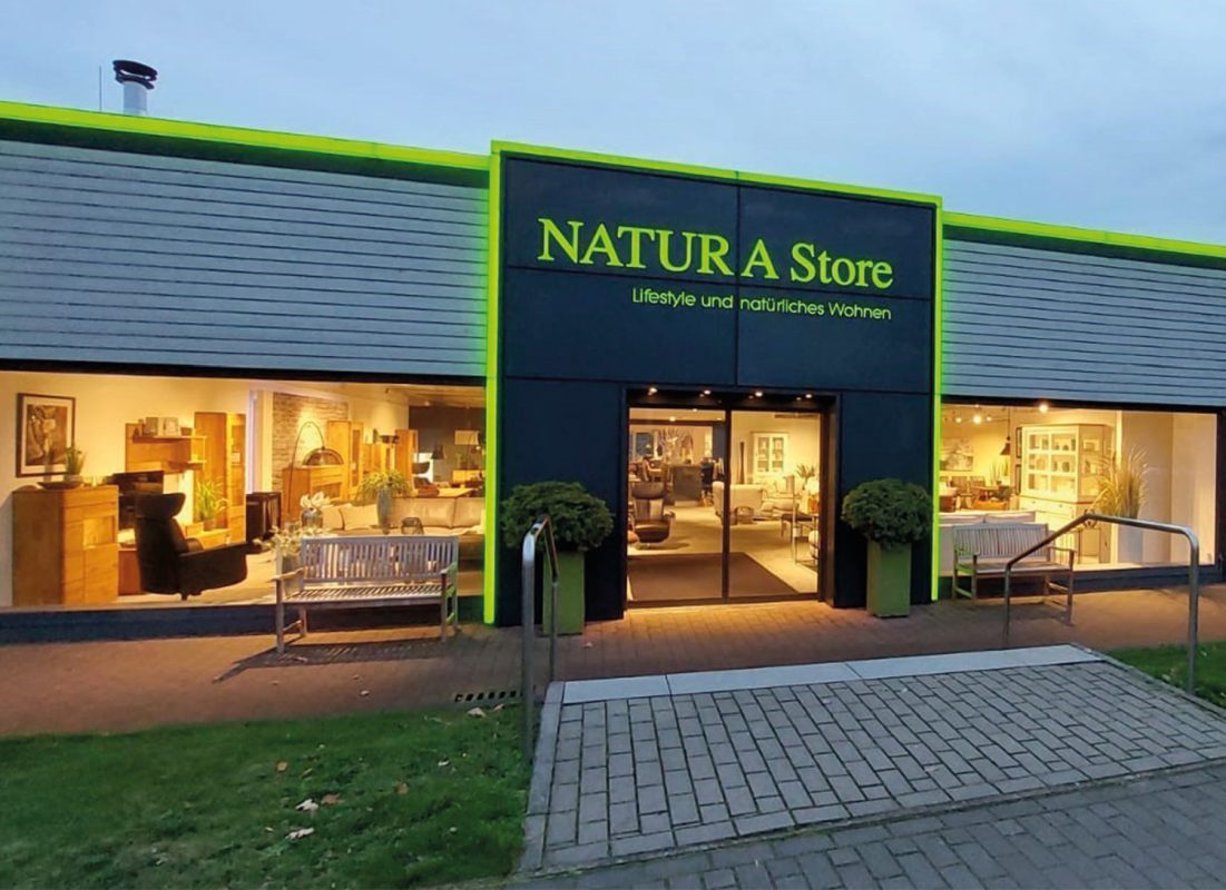 Eingangsbereich mit Schild NATURA Store, Schaufenstern, Bänken und beleuchtetem Eingang