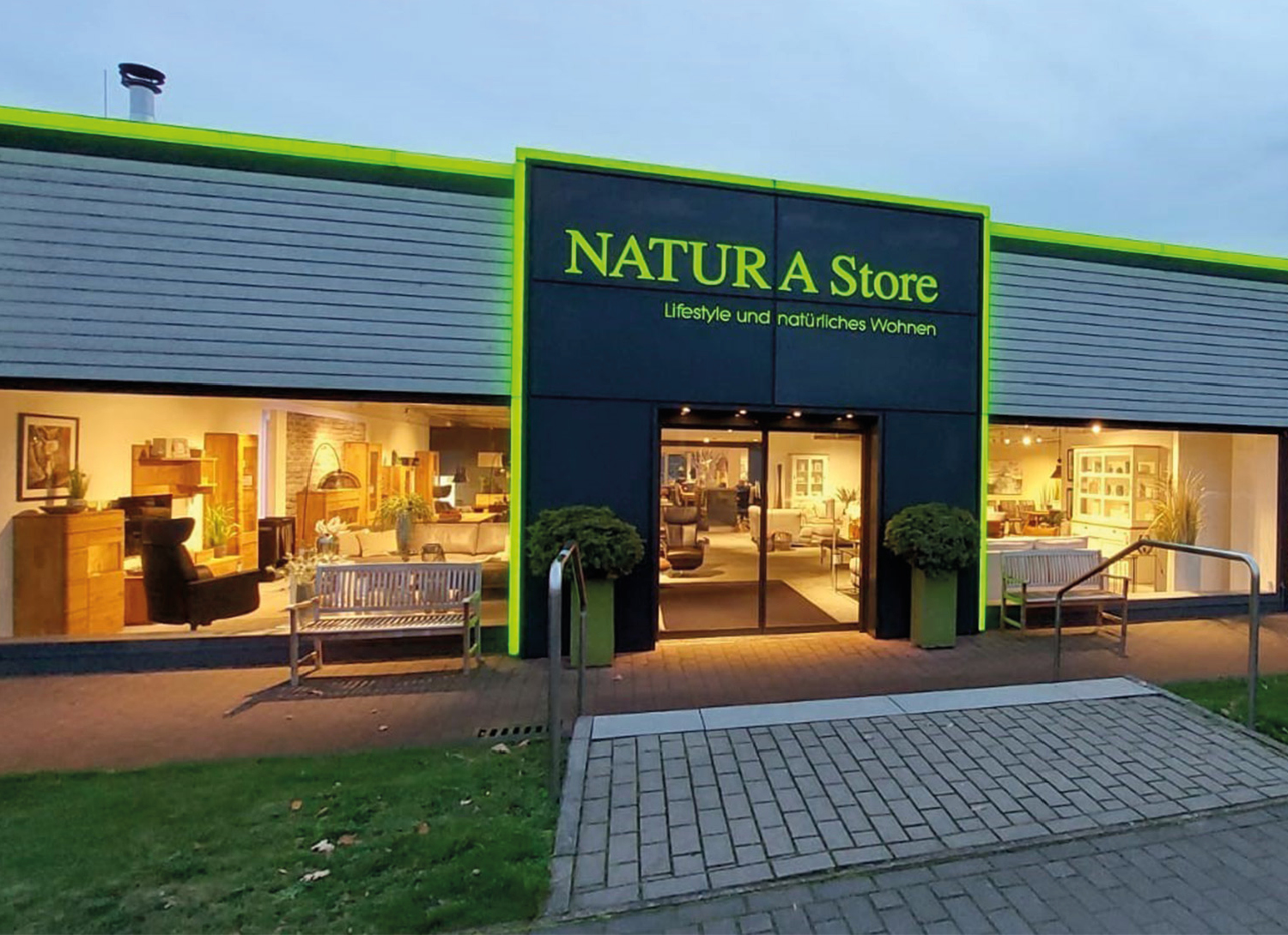 Eingangsbereich mit Schild NATURA Store, Schaufenstern, Bänken und beleuchtetem Eingang