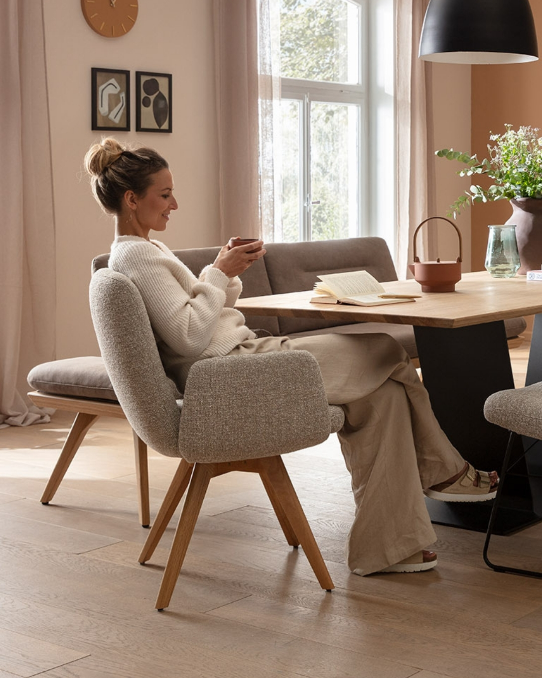 Frau in bequemer Kleidung sitzt mit einer Tasse in der Hand an einem Holztisch in einem modernen Schlafzimmer.
